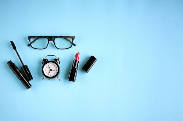 Gafas negras, despertador, rímel y pomada roja sobre fondo azul composición . — Foto de Stock