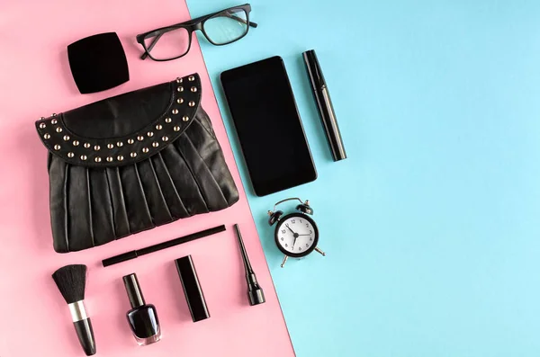 Black leather bag with nail polish, pomade, mascara, alarm clock and eye shadows on pink and blue background composition. — Stock Photo, Image