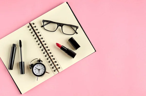 Gafas negras, despertador, bloc de notas de papel, rimel y pomada roja sobre fondo rosa composición . — Foto de Stock