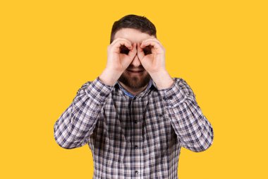 Sakallı bir adam gülümsüyor, elleriyle dürbün yapıyor, turuncu arka planda soyutlanmış. Kameraya bakıyorum..