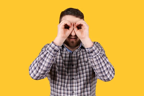 Sakallı bir adam gülümsüyor, elleriyle dürbün yapıyor, turuncu arka planda soyutlanmış. Kameraya bakıyorum..