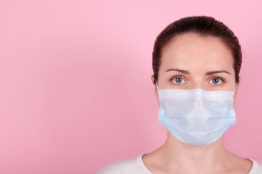Tıbbi maskeli esmer bir kızın portresi kameraya bakar. Pembe arka planda izole edilmiş. Boşluğu kopyala.