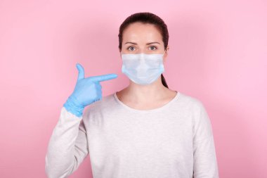 Tıbbi maskeli esmer bir kızın portresi ve yüzünde parmak olan mavi lastik eldivenler. Pembe arka planda izole edilmiş.