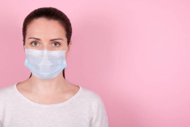 Tıbbi maskeli esmer bir kızın portresi kameraya bakar. Pembe arka planda izole edilmiş. Boşluğu kopyala.