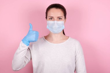 Tıbbi maske takmış esmer bir kızın portresi ve mavi lastik eldivenler iyi gösteriyor. Pembe arka planda izole edilmiş.