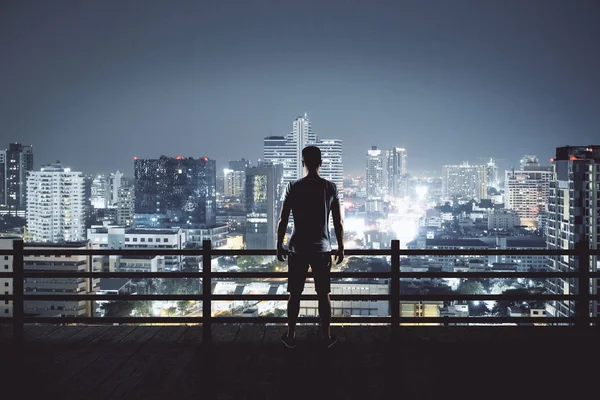 Back view of young man on rooftop looking at illuminated night city. Research and employment concept