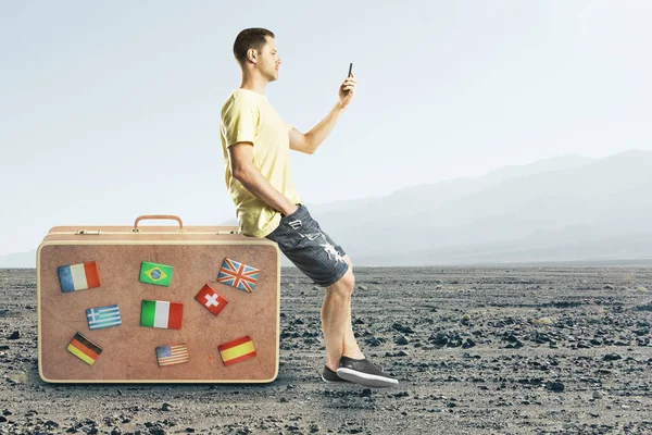 美しい風景の背景にスーツケースの上に座ってのスマート フォンを持つハンサムな欧州人の側面図です 旅行やソーシャル メディアの概念 — ストック写真