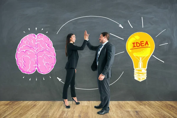 Businessman and woman giving each other hi-five on chalkboard background with brain and lamp sketch. Teamwork, brainstorm and idea concept