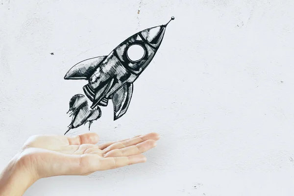 Hand holding creative rocket sketch on concrete wall background. Startup and business concept