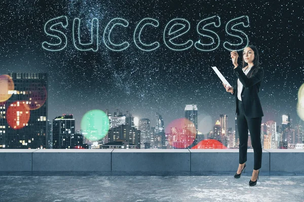 Young businesswoman writing abstract success word on concrete rooftop background with blurry night city view. Successful worker concept