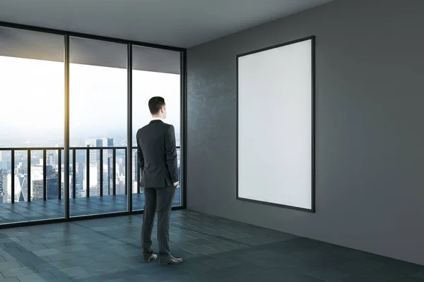 Uomo d'affari guardando finto poster bianco sulla parete scura in camera moderna . — Foto Stock