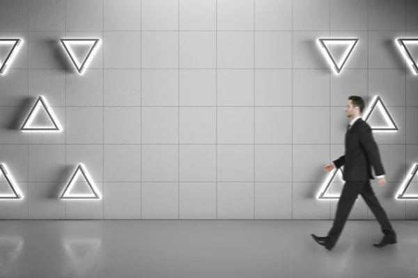 Businessman walking on concrete floor along blank grey wall with triangle lights, mock up.