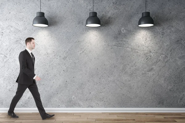 Businessman walking in gallery interior with three ceiling lamps and blank concrete wall. Gallery concept. Mock up.