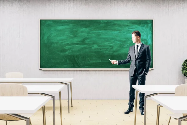 Businessman standing in modern classroom with empty chalkboard, furniture and daylight. Education and school concept. Mock up,