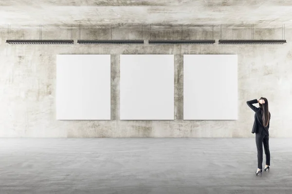 Businesswoman in gallery interior looking on three blank posters on wall. Gallery and presentation concept.  Mock up,