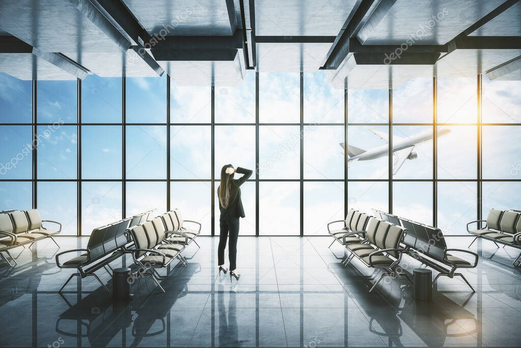 Businesswoman standing in modern airport interior with flying by airplane and sunlight view in panoramic window.