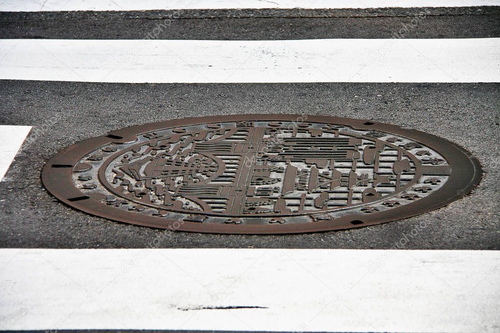 Osaka, Kansai region, Japan - September 26, 2009 - Manhole cover in Osaka engraved with a ship on Dotonbori canal and Osaka castle as symbol of an important landmark, in Zebra crosswalk background.
