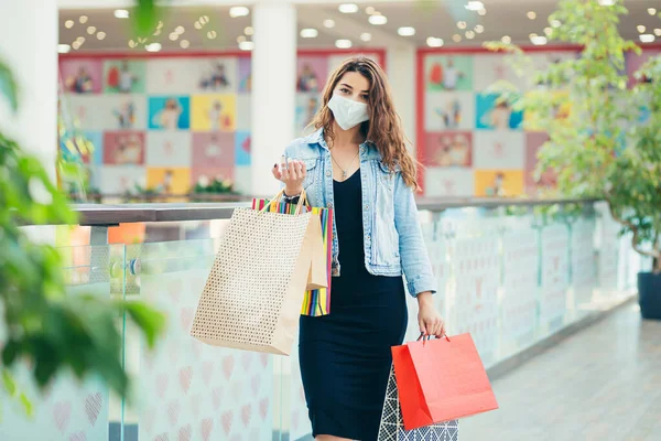 Happy young woman in medical mask walking at mall with lots of shopping bags in hands. Concept of healthcare and prevention from coronavirus.