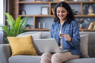 Hintli genç bir kadın evinde kanepede oturuyor, dizüstü bilgisayarını kucağında tutuyor ve elinde bir kredi kartı kullanıyor..