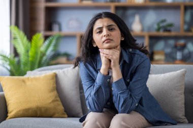 Üzgün ve endişeli genç bir Müslüman kadının portresi. Evdeki kanepede oturuyor, ellerini kavuşturuyor ve üzgün bir şekilde kameraya bakıyor..