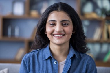 Kapalı alanda kot gömlekli, ayakta kameraya gülümseyen ve gülümseyen genç bir Hintli kadının yakın plan portresi..