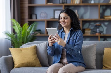 Smiling young Indian woman sitting on the sofa at home and using her tablet.