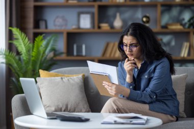 Serious young Indian woman sitting on sofa at home and counting bills, looking at receipts and documents.