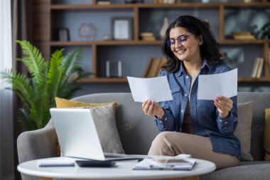 Smiling young Indian woman sitting at home on the couch at a table with a laptop and working with documents and receipts.