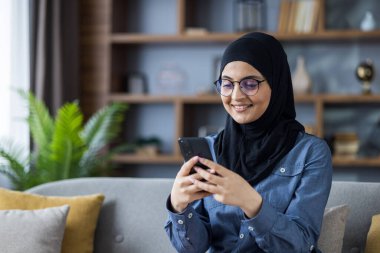 Smiling young Islamic woman in hijab and glasses sitting on sofa at home and using mobile phone.