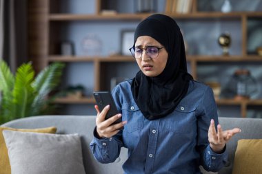 Worried young Muslim woman in hijab sitting on sofa at home, looking at mobile phone screen and spreading her hands in frustration.