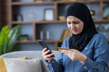 Worried Muslim young woman in hijab sitting on the sofa at home, holding a phone in her hand and looking at a credit card with sadness.
