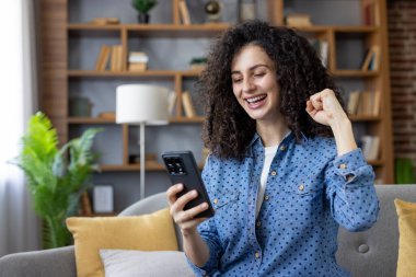 Ateşli, kıvırcık saçlı bir kadın rahat bir koltukta oturuyor, sevinçle yumruğunu kaldırıyor, akıllı telefonuna bakıyor, heyecan ve başarı saçıyor.