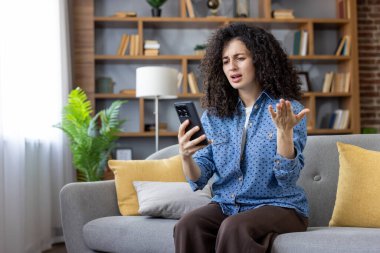 Genç bir kadın, rahat bir oturma odasında kanepede oturmuş, biriyle tartışırken akıllı telefonuyla bir video görüşmesi yapıyor, konuşma sırasında hayal kırıklığını ve asabiyeti dile getiriyor.