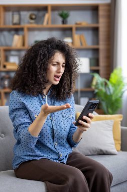 Oturma odasındaki rahat bir kanepede oturan kıvırcık saçlı genç bir kadın ateşli bir video görüşmesi yapıyor. Hayal kırıklığını ve el hareketleriyle ifade ediyor.