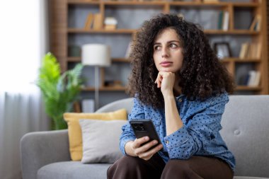 Düşünceli kadın serbest çalışan ev ofisinden uzak bir iş projesi üzerinde kafa yoruyor, cep telefonunu elinde tutuyor ve başka yere bakıyor.