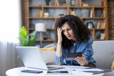 Endişeli genç kadın oturma odasında bir kanepede oturuyor, hesap makinesine bakarken başını tutuyor ve ev bütçesini ve kişisel mali durumunu kontrol etmek için dizüstü bilgisayar kullanıyor.