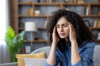Stresli kadın kendini sağlıksız hissediyor, şakaklara dokunuyor, migrenden muzdarip, kafasında ağrı olan, oturma odasındaki kanepede oturan, baş dönmesi yaşayan, kronik baş ağrısı çeken