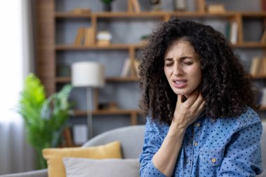 Boynuna dokunurken boğaz ağrısı çeken genç bir kadın evdeki bir kanepede oturuyor. Bademcik iltihabı ya da gırtlak iltihabı belirtileri gösteren bir soğuk algınlığı belirtisi gösteriyor.