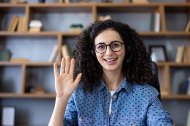 Dost canlısı, kulaklık takan ve gözlük takan kadın destek çalışanı merhaba diyor, online görüşme sırasında müşteriyi selamlıyor, video konferans çağrısına başlıyor, müşteriye yardım ediyor.