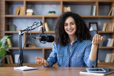 Neşeli bir kadın podcaster, içerik yaratımı için profesyonel bir mikrofon ve dijital tablet kullanarak, bir ev stüdyosunda bir ses kaydı oturumunda el kol hareketi yapıyor.