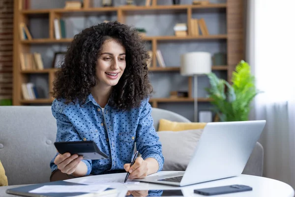 Evde çalışan, hesaplayan ve not alan genç bir kadın, finansal raporları gözden geçirmek için bilgisayarını kullanıyor, iç finansmanı verimli ve rahat bir şekilde yönetiyor.