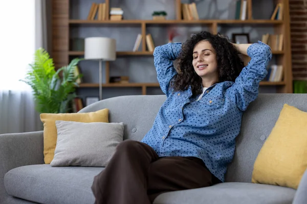 Genç bir kadın rahat koltuğunda bir anlık huzur ve rahatlığın tadını çıkarıyor, elleri başının arkasında, gözleri kapalı, yüzünde huzurlu bir gülümseme, oturma odasının rahat atmosferiyle çevrili.