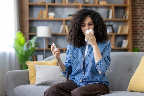 Evdeki kanepede oturan hasta genç bir kadın, burun spreyi tutarken burnunu mendille siliyor, soğuk algınlığı veya grip ile boğuşuyor.