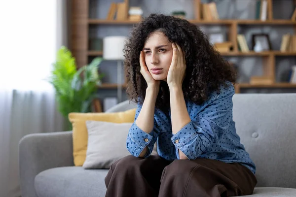 Evdeki kanepede oturan üzgün kadın, baş ağrısı, yorgunluk, stres ve yalnızlık hissederken şakaklarına dokunuyor.