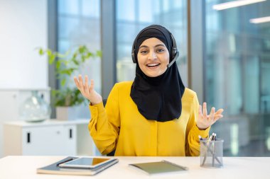 Dost canlısı bayan müşteri hizmetleri temsilcisi kulaklık ve tesettür takıyor ve modern ofiste masasında çalışırken jestler kullanarak bir şeyler anlatıyor.