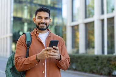 Genç Latin üniversite öğrencisi kampüs binasının yanında akıllı telefon uygulaması kullanarak gülümsüyor, günlük kıyafetler giyiyor ve yeşil bir sırt çantası taşıyor, açık havada keyifli bir gün geçiriyor.