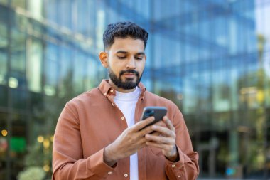 Modern bir ofis binasının önünde dururken akıllı telefon tutan genç işadamı, İnternet 'i karıştırıyor ve kentsel bir ortamda müşterilerin veya iş arkadaşlarının mesajlarını okuyor