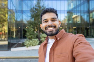 Genç işadamı, modern bir ofis binasının önünde selfie çekerken gülümsüyor. Mesleki hayatında bir başarı ve güven anı yakalıyor.
