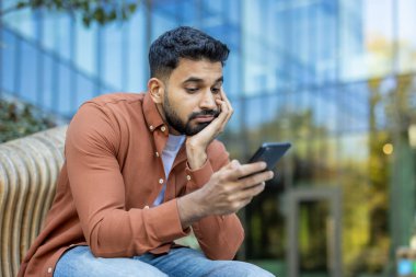 Genç işadamı bir bankta oturuyor, akıllı telefonuyla kayarken sıkılmış görünüyor. Pürüzsüz, modern bir cam binanın önünde yüzünü eline dayadı.