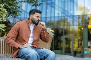 Şehirdeki bir bankta oturan sakallı adam, öksürüyor ve yumruğuyla ağzını kapatıyor, hastalık ya da alerjiden muzdarip.